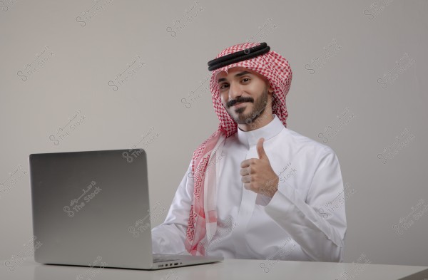 A portrait of a man sitting in front of a laptop, giving a thumbs-up with a smile. The man is wearing a white thobe and a red checkered ghutra with a black agal, indicating traditional Saudi attire.