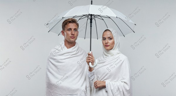 A portrait of a man and woman standing under a white umbrella. The man is wearing traditional white Ihram clothing, and the woman is wearing a white hijab with a white dress. The background is a light gray, presenting a calm and serene atmosphere.