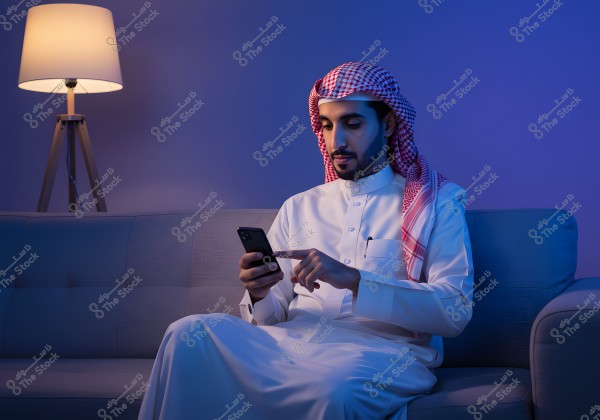 Portrait of a man sitting on a sofa, wearing a white thobe and a red and white checkered ghutra with an igal, suggesting a possible Saudi origin. He is using his smartphone. In the background, there is a modern floor lamp emitting warm light, and the ambiance is enhanced by soft blue lighting, adding a serene touch to the scene.