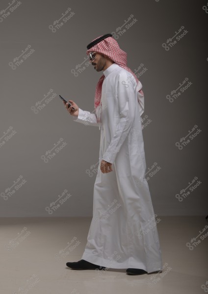 Image of a man wearing traditional Gulf attire, consisting of a white thobe, a red checkered headscarf (ghutra), and a black agal. The man is holding a mobile phone and looking at it while walking. The background is gray and neutral, highlighting the details of the attire.