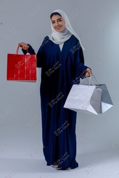 An image of a woman wearing a dark-colored abaya and a white hijab, carrying red and white shopping bags in her hands. She appears smiling against a neutral gray background.