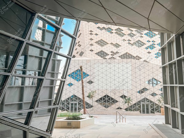 Image of a modern building with a unique architectural design featuring tilted glass windows. In the background, another building showcases a geometric design with triangular and square shapes. The foreground includes an entrance with stairs and a few tall trees.