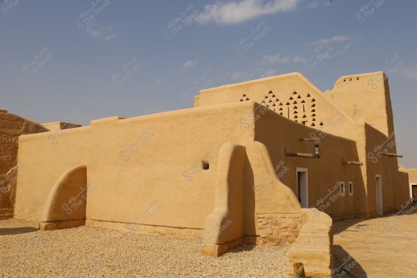View of a traditional mud-brick building in a desert area, reflecting the Najdi architectural style commonly found in Saudi Arabia. The building features thick walls and small triangular ventilation windows. The sky is clear blue with a few light clouds.