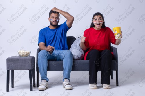 Portrait of a Saudi young man and woman watching a match and cheering their favorite teams cheerfully while having a snack on a blue background, the World Cup.