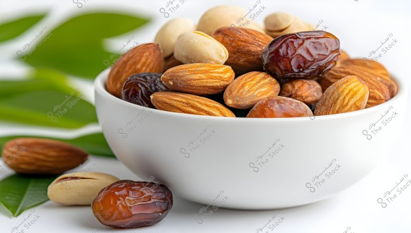 An image of a white bowl filled with a variety of nuts and dates, with green leaves in the background. The bowl contains almonds, pistachios, and shiny brown dates.