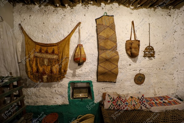 The image shows a wall adorned with traditional old tools. On the white wall with a wooden shelf at the top, there is a brown wall tapestry, a traditional pouch, and a woven straw bag. In the center, an old radio is placed in a niche in the wall surrounded by green paint. Below the wall, there\'s a brown wooden bench with fabric cushions decorated with colorful geometric patterns.