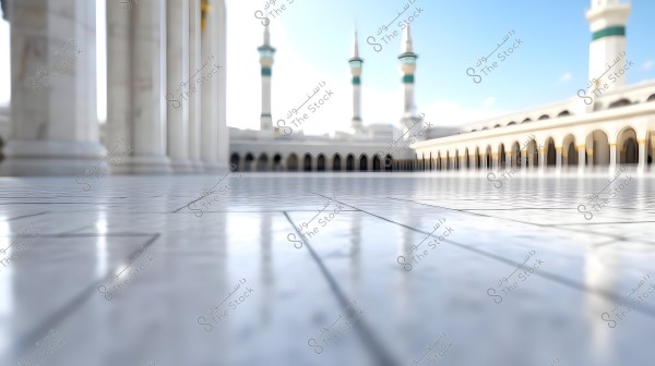 The image shows a large open courtyard with white marble flooring, featuring massive columns on the sides and prominent minarets in the background under a clear blue sky. The architectural design suggests it is part of an Islamic mosque in Saudi Arabia.