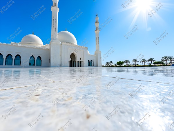 View of a large mosque building featuring a white dome, a secondary dome, and tall minarets, with a clear blue sky and bright sun shining above. The foreground is covered in shiny marble reflecting the light. Palm trees and greenery are visible in the background.