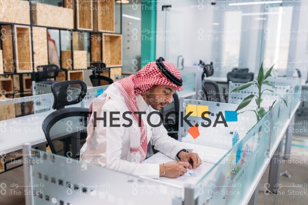 رجل سعودي يرتدي السعودي التقليدي يجلس في المكتب بينما يدون ملاحظته، موظف أثناء العمل في الشركة