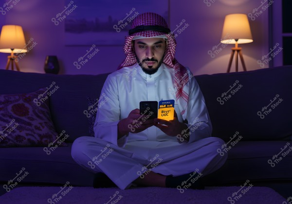 A man sitting on a couch in a room with dim night lighting, wearing traditional attire consisting of a thobe and a red shemagh. He appears to be from an Arab country, possibly Saudi Arabia. He is holding a mobile phone and a card, with the phone\'s light illuminating his face. Behind him are lit lamps and a painting on the wall.