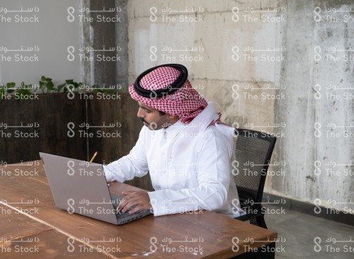 رجل سعودي يرتدي الزي السعودي التقليدي يستخدم الابتوب ويدون الملاحظات في دفتر أثناء جلوسه في مكتبه، موظف يعمل في المكتب