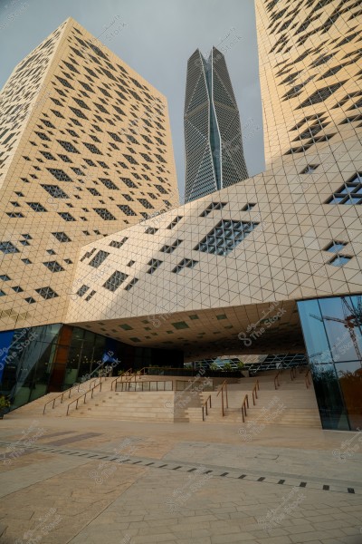 A view of several modern towers in a cityscape in Riyadh, Saudi Arabia. The exterior design features interlocking geometric patterns and repeated small windows. The central tower is the tallest with a unique prismatic design. Stairs lead up to the entrance below, with construction cranes visible in the background.