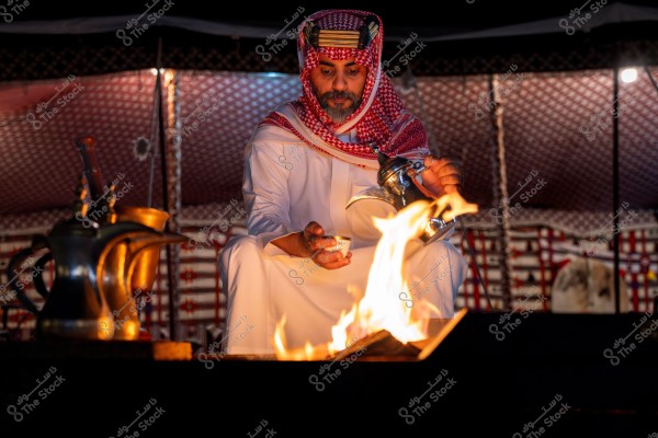 A man dressed in traditional Saudi attire with a thobe and shemagh, sitting inside a decorated tent. He is pouring coffee from an Arabic dallah into a small cup in front of a burning fire. The tent is adorned with traditional patterns, and there are two dallahs beside the man.