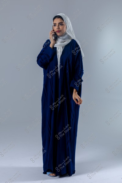 A portrait of a woman wearing a dark blue abaya and a white hijab with dotted patterns, seemingly speaking on a mobile phone. She stands in a relaxed pose against a grey background. A part of her open-toed blue sandals is visible.