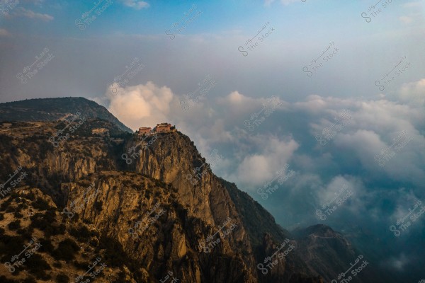 A natural landscape of a high mountain in the Asir region, Saudi Arabia, featuring a building perched on the edge of the mountain with thick clouds in the background. The mountain is covered with scattered vegetation, extending into the horizon where the clouds meet its peaks.
