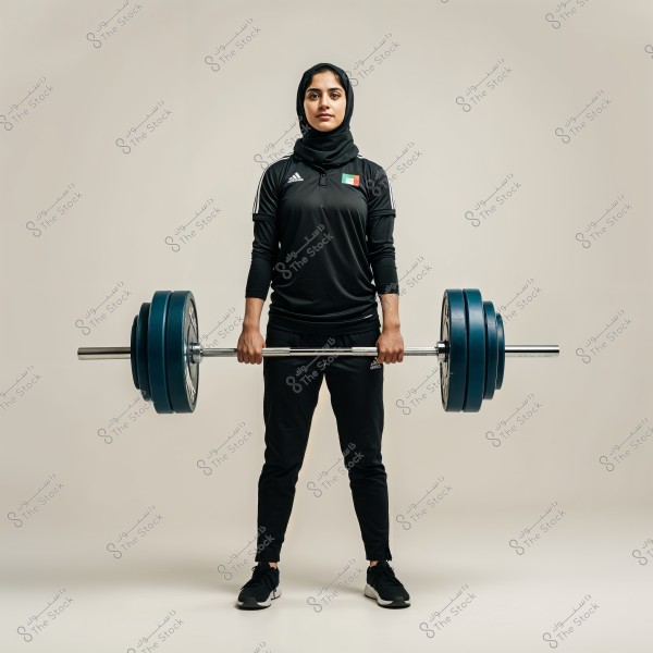 An image of a woman wearing a black sports outfit with a hijab, holding a barbell steadily. The UAE flag is visible on her shirt. The background is simple with a neutral color, highlighting her focus and determination.