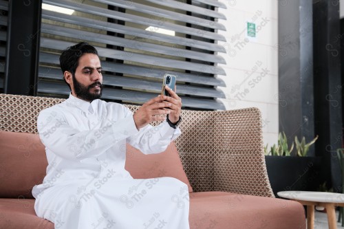 رجل سعودي يرتدي الثوب السعودي يجلس في غرفة الإجتماعات يحمل الهاتف المحمول بينما يقوم بتعابير تدل على الإنزعاج