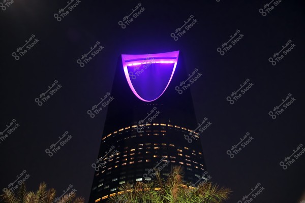 Night view of the Kingdom Tower in Riyadh, Saudi Arabia. The tower is prominently lit at the top with a glowing purple arc. Windows are partially lit, and palm trees can be seen at the base.