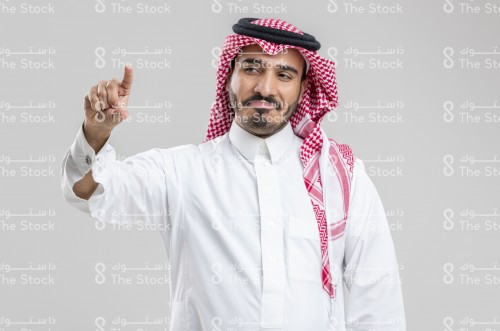 بورتريه رجل سعودي يرتدي الزي السعودي التقليدي  يشير بأصابعه السبابة والإبهام يسار الصورة