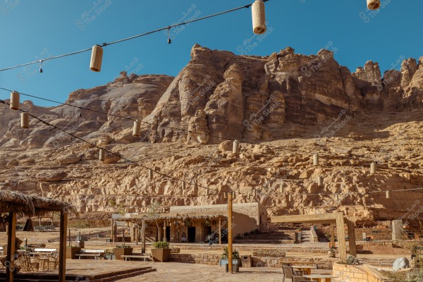 A natural landscape showing a desert area with towering rocky mountains in the background under a clear blue sky. In the foreground, there is an outdoor seating area with tables and chairs, and a small rustic building covered with mud. There are also hanging lights on wires stretched across the image.