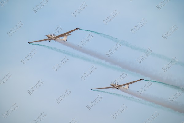 Two white airplanes flying side by side in a blue sky, leaving trails of bluish-white smoke behind them. The airplanes are flying in a synchronized formation.