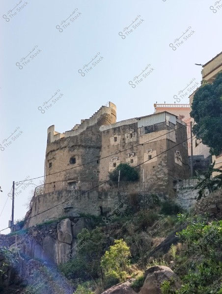 The image shows a castle or traditional stone structure on top of a hill, consisting of several stories with thick walls and small windows. The structure is surrounded by a stone fence and rises above natural rocks and vegetation. The sky is clear in the background.