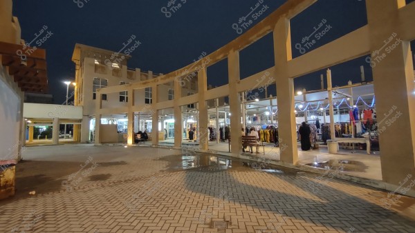 A nighttime scene of an open-air market featuring a large building with traditional Arabic architectural lines. Stalls displaying clothes in various colors are seen under the night lights. People are shopping and wearing traditional attire, and part of the outside courtyard is visible with stone paving and a light post.