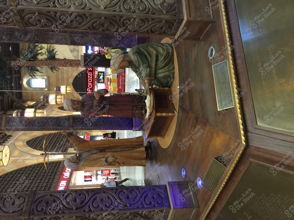 Statues of historical figures wearing turbans and traditional clothing, displayed in a scene within a shopping mall. There is a decorative wooden structure surrounding the statues, with a background of ornate windows and illuminated palm trees, as well as store signs.