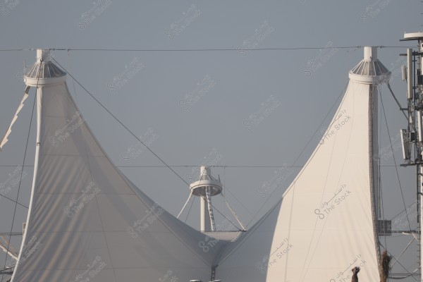The image features large tent-like architectural structures made of white fabric material. They are centered by thin supporting poles and connected by cables, forming part of a modern architectural design. Communication tower equipment is visible on the right side under a clear blue sky.