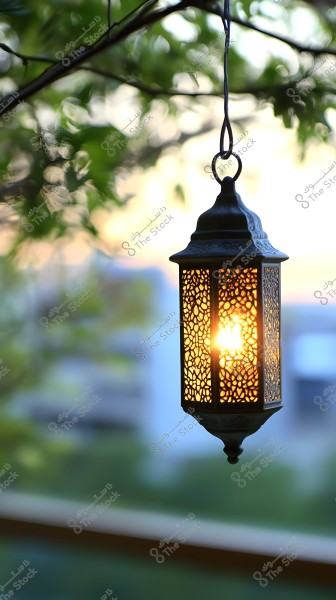 An ornate metal lantern hanging from a tree branch, glowing with a warm yellow light. The lantern has an intricate eastern design, allowing light to filter through. The background features green leaf details with a bokeh effect, focusing attention on the lantern.