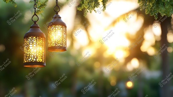 Two decorative metal lanterns hanging from tree branches, emitting a warm glow in an outdoor natural setting. The surrounding lights illuminate the scene, which includes trees and other foliage in the background, lit by sunlight during sunset.