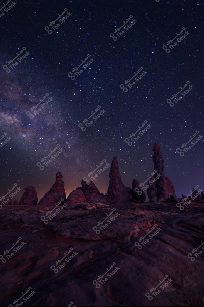 A nighttime image showcasing a star-filled sky with prominent rock formations in the foreground. The rocks rise dramatically over the sandy terrain, while the horizon is bathed in faint light. The colors range from deep blues to purples.
