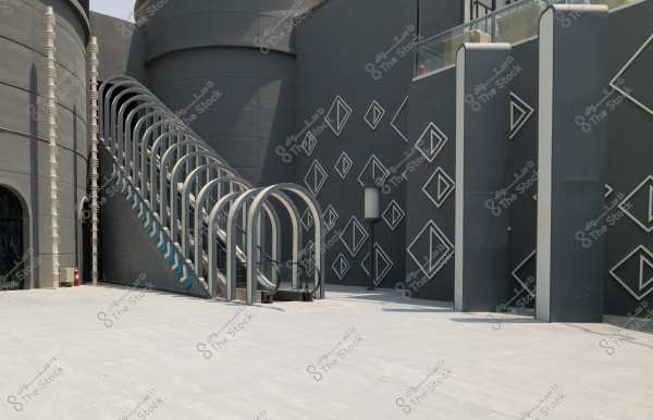 Image of a modern building with a contemporary design, featuring dark walls adorned with repetitive diamond-shaped geometric patterns. There is an escalator enclosed by a metal structure with a series of arches. A large cylindrical structure is next to the building, and the upper platform includes a glass barrier. A vertical metal column and geometric decorations add aesthetics to the space.
