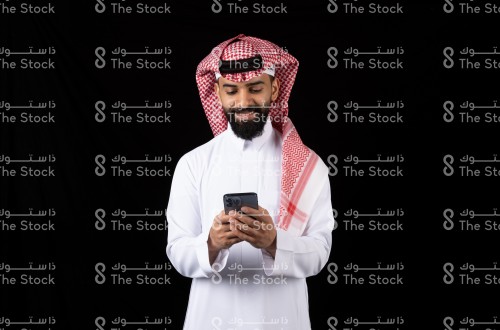 بورتريه رجل سعودي  يقف على خلفية سوداء يرتدي الثوب يحمل جوال وينظر اليه وهو مبتسم، رجل سعودي يحمل جوال بيديه مع خلفية سوداء