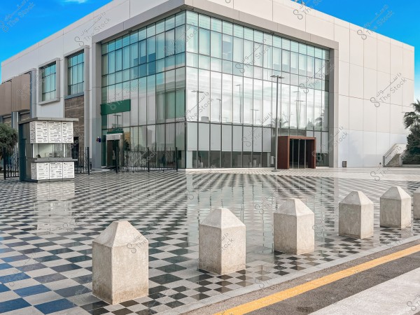 A modern building with a large glass design and a front courtyard covered with patterned black and white tiles. A small guard kiosk is near the main entrance, surrounded by metal gates and stone bollards, with green trees visible to the right and a clear blue sky in the background.
