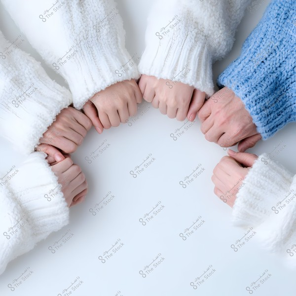 An image showing a group of hands gathered in a circular arrangement on a white surface. The hands are clenched, wearing knitted sweaters in white and light blue. The image focuses on the knitted sleeves with different textures, suggesting warmth.