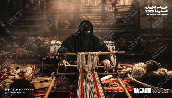 A photo of a woman wearing a niqab and traditional attire, sitting in front of a handloom weaving a rug. The background is adorned with textiles featuring traditional patterns. Baskets containing yarn and wool are beside her. The image represents traditional handicrafts and includes text \"The Year of Handicrafts 2025\" along with the Saudi Ministry of Culture logo.