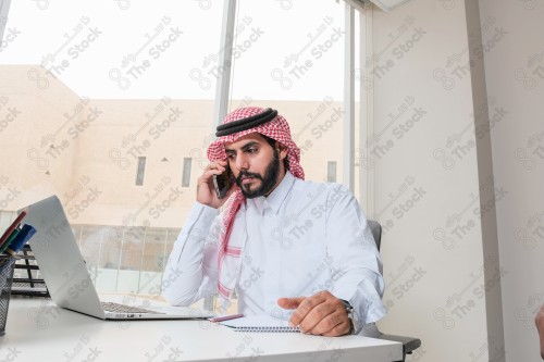 رجل سعودي يرتدي الزي السعودي التقليدي يعمل في مكتب بواجهة زجاجية ويستخدم قلم رصاص لتدوين الملاحظات