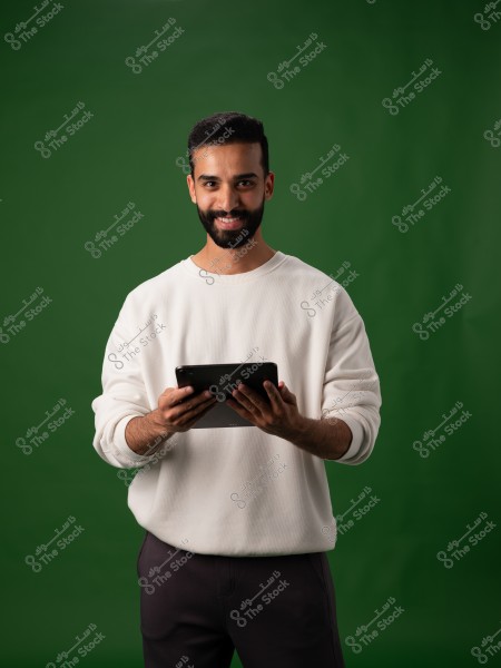 A portrait of a man with a short beard wearing a white sweater and dark pants, standing against a green background. He is holding a tablet with both hands and looking at the camera with a slight smile.