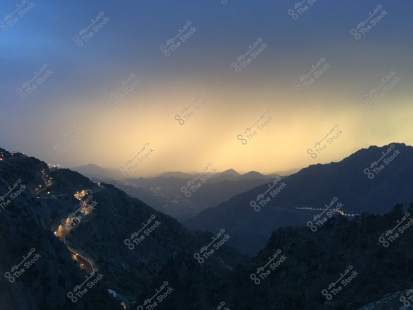 Stunning natural view of fog-covered mountains in the evening. A winding road with illuminated walkways stretches through the highlands, while the sky glows with vibrant yellow and orange hues at sunset, surrounded by layered mountains in the horizon.