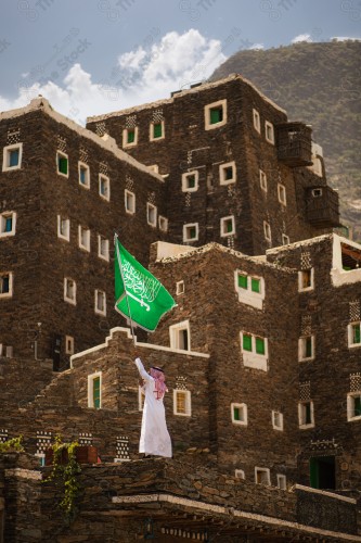 A Saudi man in traditional Saudi dress holds the Saudi flag over a building in Rijal Almaa village during the day in the Asir region. A Saudi man holds the Saudi flag in celebration of the National Day.