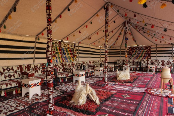 The image shows a traditional Bedouin tent beautifully decorated. The tent features bright colors and geometric patterns on the blankets and cushions. The interior walls are made of striped fabric in white and black colors. The ceiling is supported by poles adorned with colorful patterns, and multicolored lights and lanterns hang from the ceiling. The floor is covered with red patterned rugs with traditional designs. There are pottery jars and low seating furniture.