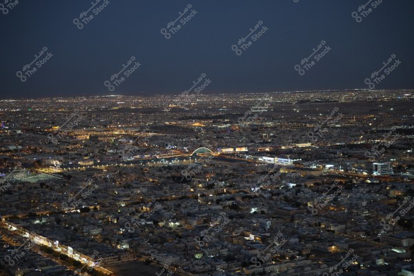منظر جوي لمدينة كبيرة في الليل تظهر أنوار المباني والشوارع متلألئة وسط الظلام. يظهر في الصورة جسر مضاء بألوان زاهية يتقاطع مع المدينة. يمكن رؤية الأفق المتناثر بأنوار المدن البعيدة.