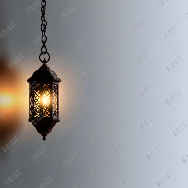 A decorative metal lantern with intricate geometric designs suspended by an iron chain. The light emanating from the lantern casts a warm glow against a calm grey background.