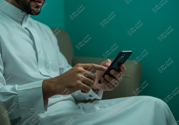 The image shows a person sitting on a green sofa, wearing a traditional white thobe, and holding a smartphone while pointing at its screen. The wall behind is dark green, creating a relaxed ambiance.