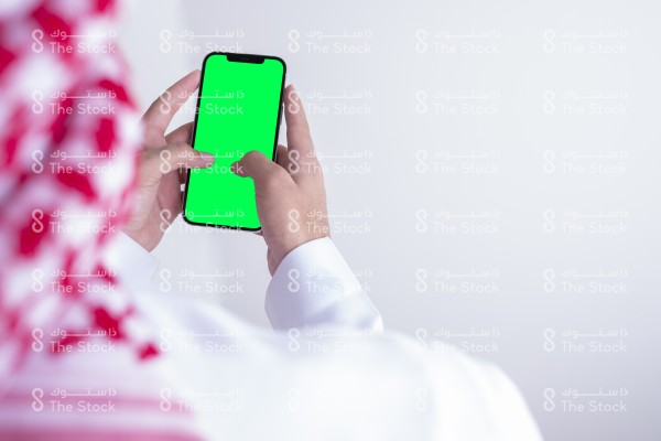 صورة ملتقطة بهاتف من الخلف لرجل سعودي يرتدي الزي السعودي التقليدي يستخدم الجوال وتظهر شاشة الجوال بوضوح