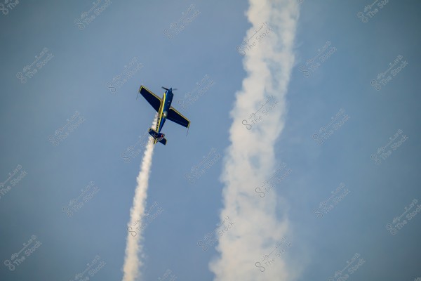 A blue aircraft with yellow accents flying at a steep angle in the blue sky, leaving a thin white smoke trail behind, with other smoke trails in the background.