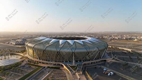 An aerial view of King Abdullah Sports City Stadium in Jeddah, Saudi Arabia. The stadium features a unique architectural design resembling a flower, with intricate details on the golden roof adorned with white geometric shapes. It is surrounded by a vast area with parking spaces and palm trees. The cityscape stretches in the background under a clear sky.