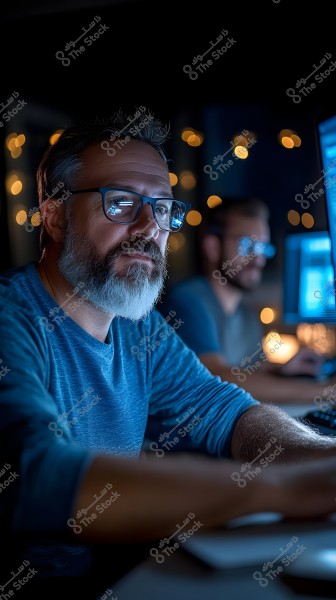 Image of a man wearing glasses and a blue shirt, seated in front of a computer in a dimly lit environment. Soft lights are visible in the background, and he appears focused on his work. Another person is sharing the screen, highlighting a collaborative nighttime work atmosphere.