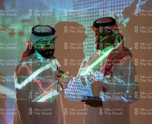 رجلان سعوديان يرتديان الزي السعودي التقليدي يمسكان بالأوراق ويتناقشان أثناء متابعة سوق الأسهم بينما تنعكس أضواء الشاشة عليهما